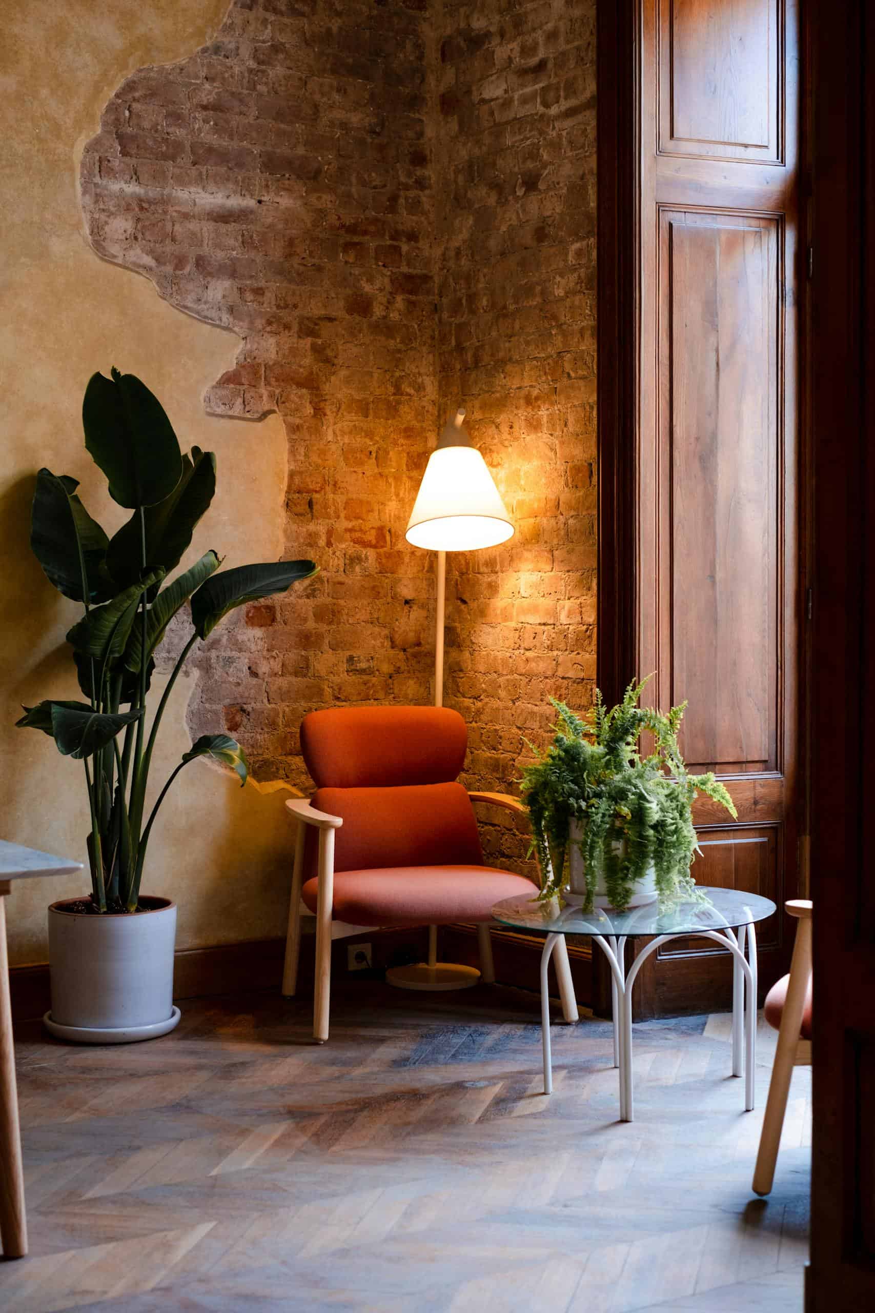 Warm living space featuring a red brick wall, armchair, glow from a lamp, and houseplants.